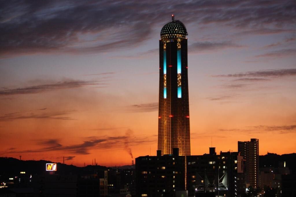 A tall, rectangular building with a dome on top, illuminated with blue and red lights, set against a sunset sky.