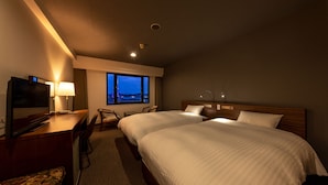 A hotel room with two beds, a desk with a TV, a chair, and a view of the city at dusk. - Plaza Hotel Shimonoseki (Shimonoseki)
