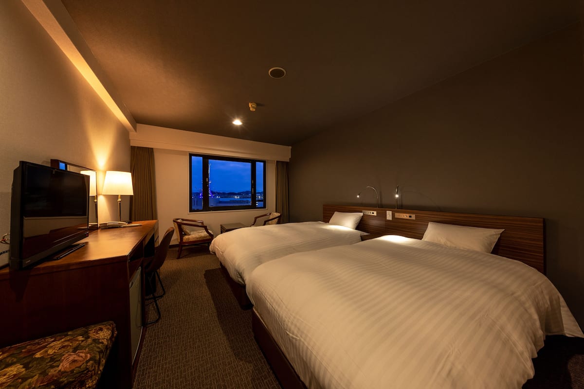 A hotel room with two beds, a desk with a TV, a chair, and a view of the city at dusk.