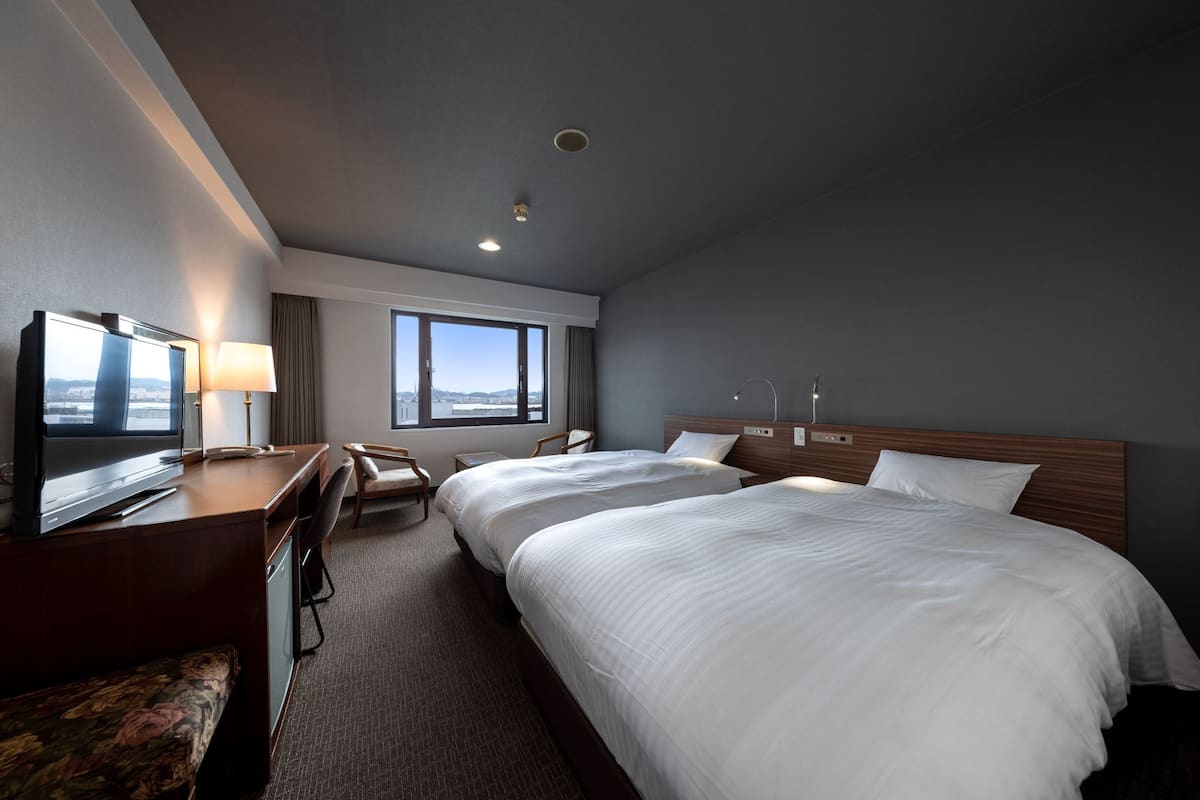 A hotel room with two beds, a desk with a TV, a chair, and a view of the city.