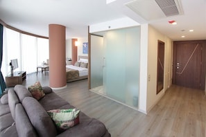 A modern hotel room with a large bed, a sofa, a glass partition, and a wooden wardrobe. - Forum Suite Hotel (Mersin)