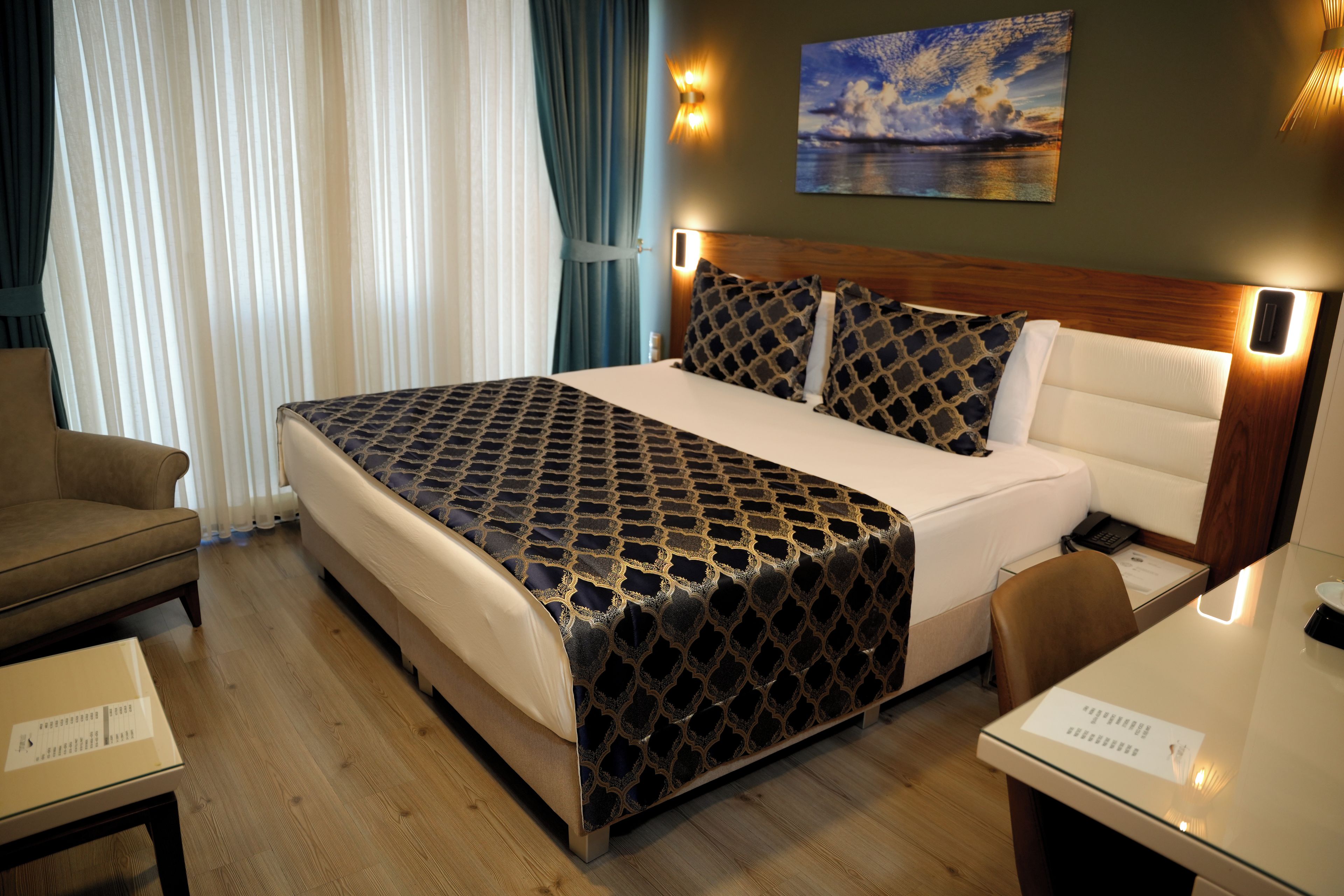 A hotel room with a large bed, a wooden headboard, a painting on the wall, and a desk with a chair.
