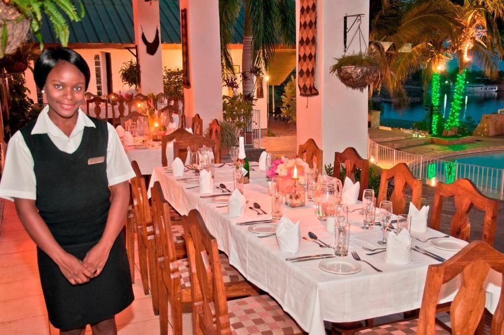 A restaurant with a long table set for dining, wooden chairs, and a pool area with illuminated plants.