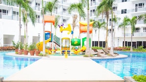 A swimming pool area with a children's play structure, palm trees, and a multi-story building in the background. - Shell Residences Apartment by Homebound (Pasay)