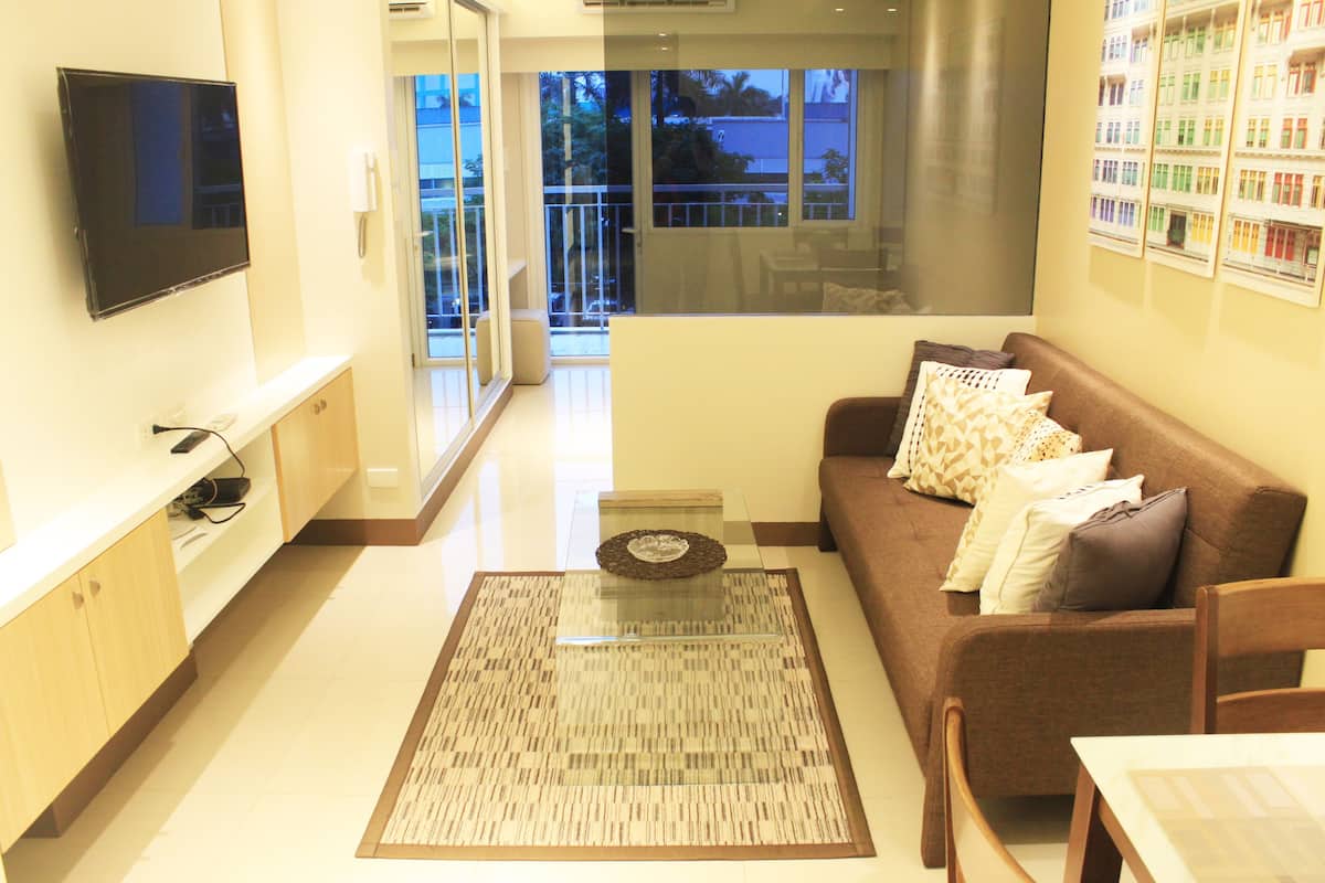 A modern living room with a flat-screen TV, a brown sofa with patterned cushions, a glass coffee table, and a dining area with a table and chairs.