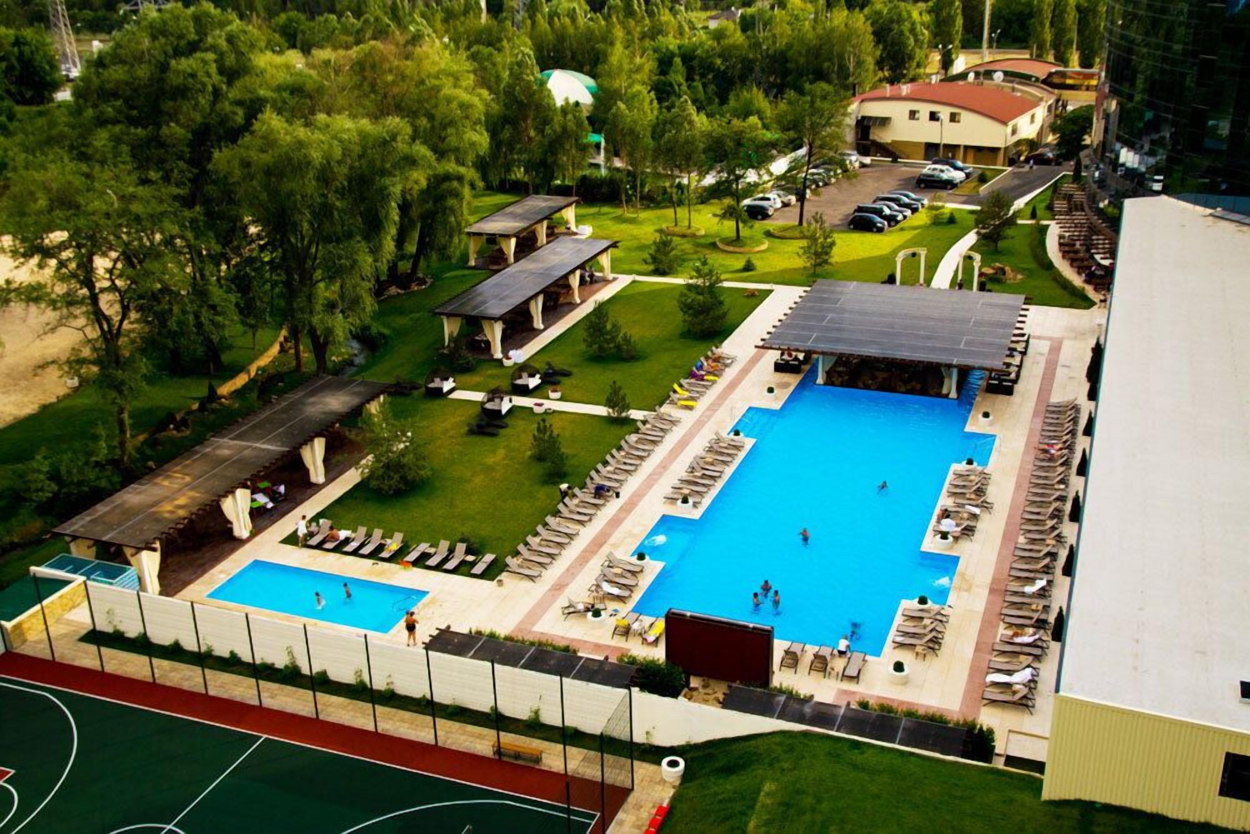 An aerial view of a resort with a large swimming pool, sun loungers, and a tennis court.