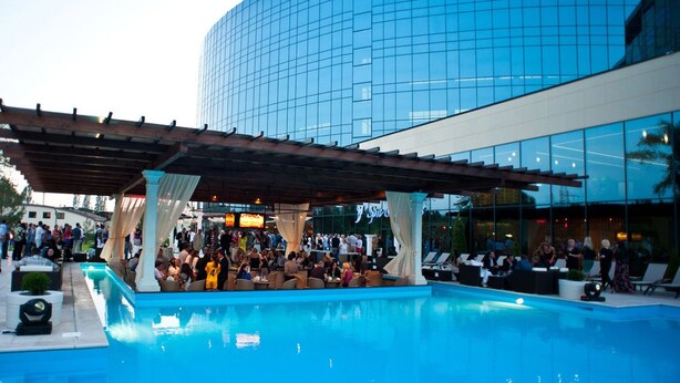 Un espace piscine doté de chaises longues et d’un coin salon abrité, situé à côté d’un bâtiment moderne aux fenêtres en verre réfléchissant.