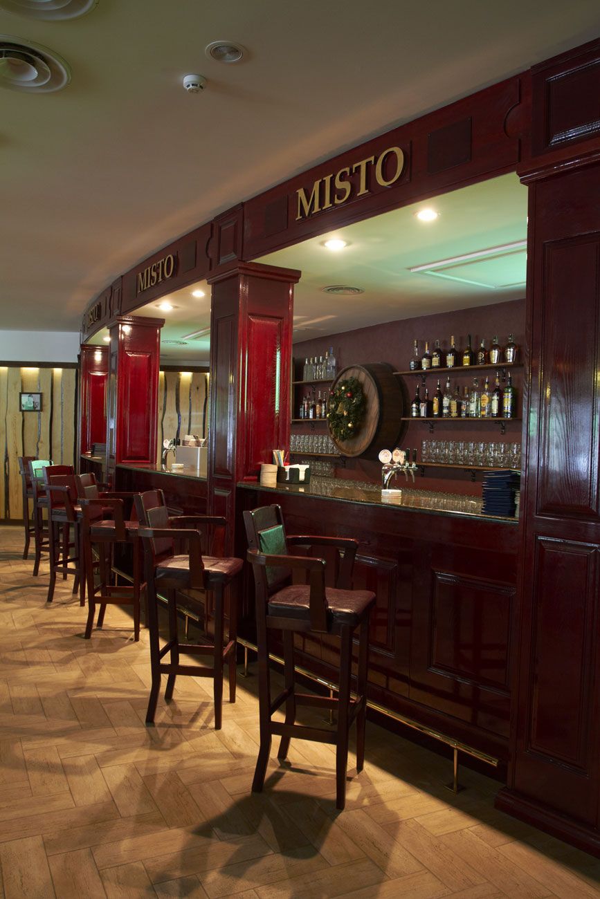 A bar area with a wooden bar counter, multiple bar stools, and shelves with bottles and glasses.