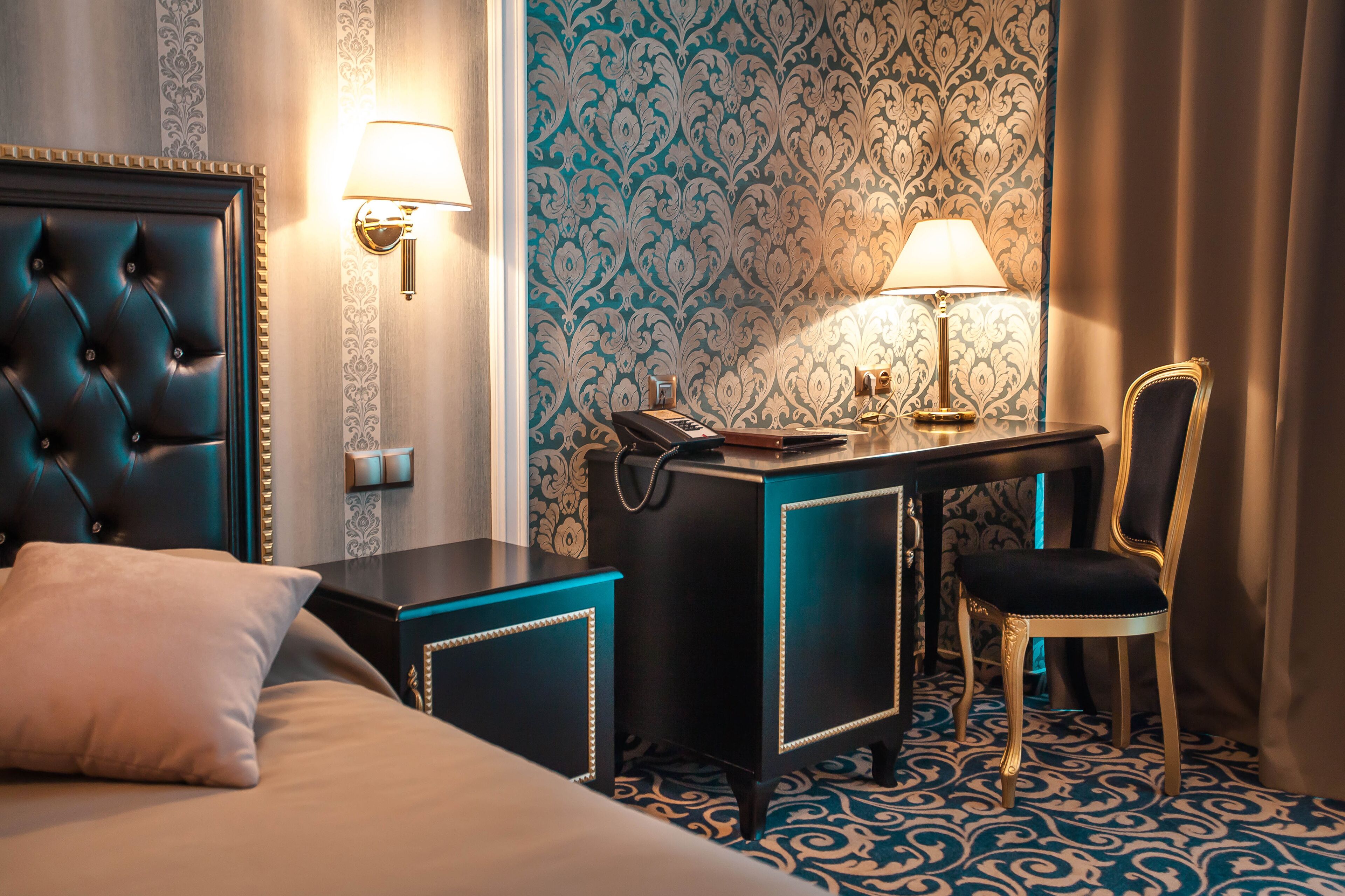 A hotel room with a tufted headboard, a desk with a lamp, a chair, and a patterned carpet.