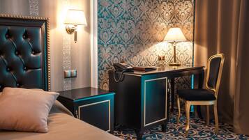 A hotel room with a tufted headboard, a desk with a lamp, a chair, and a patterned carpet.