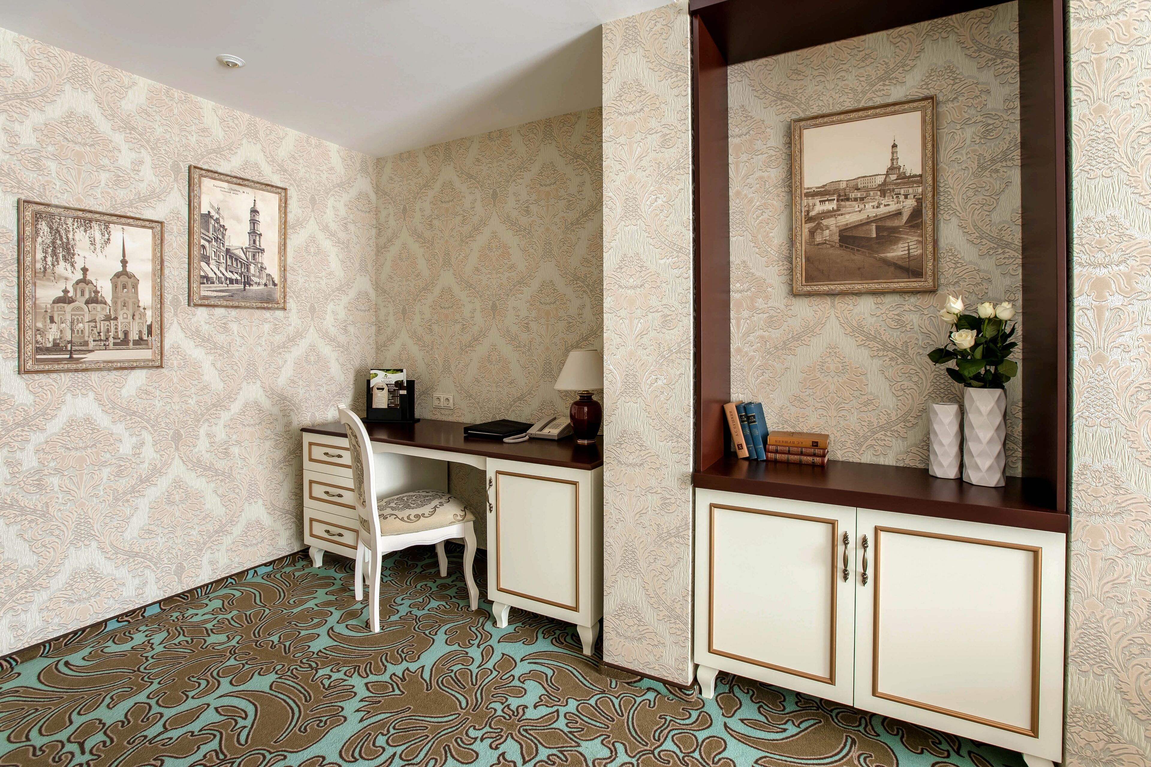 A room with patterned wallpaper, a desk with a telephone, a chair, a cabinet with books, and framed pictures on the wall.