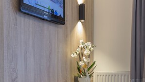 Una camera moderna con una scrivania in legno, una sedia nera, una televisione a schermo piatto montata a parete e un vaso con orchidee bianche.