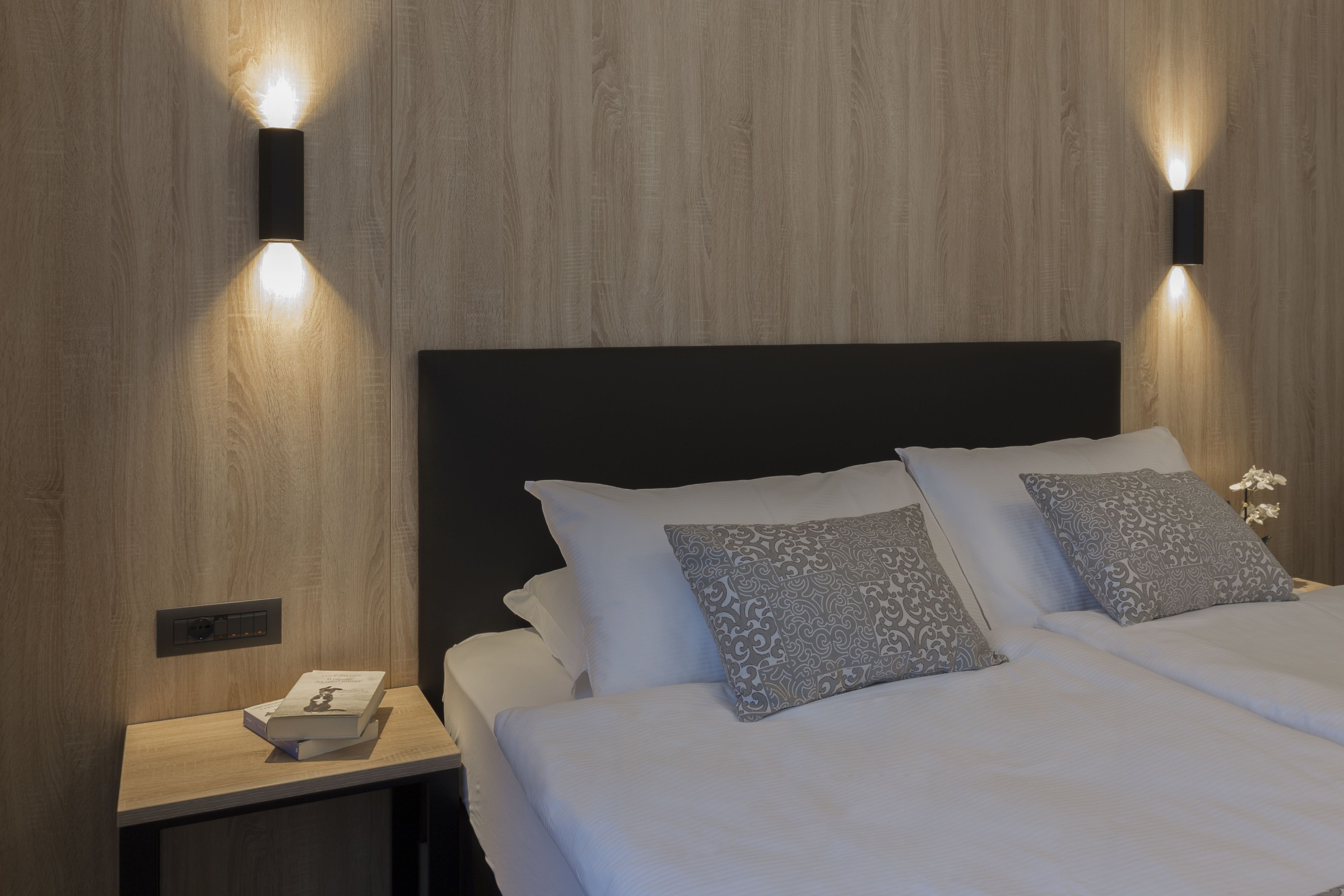 A neatly made bed with white linens and patterned pillows, a bedside table with a book, and two wall-mounted light fixtures on a wooden wall.