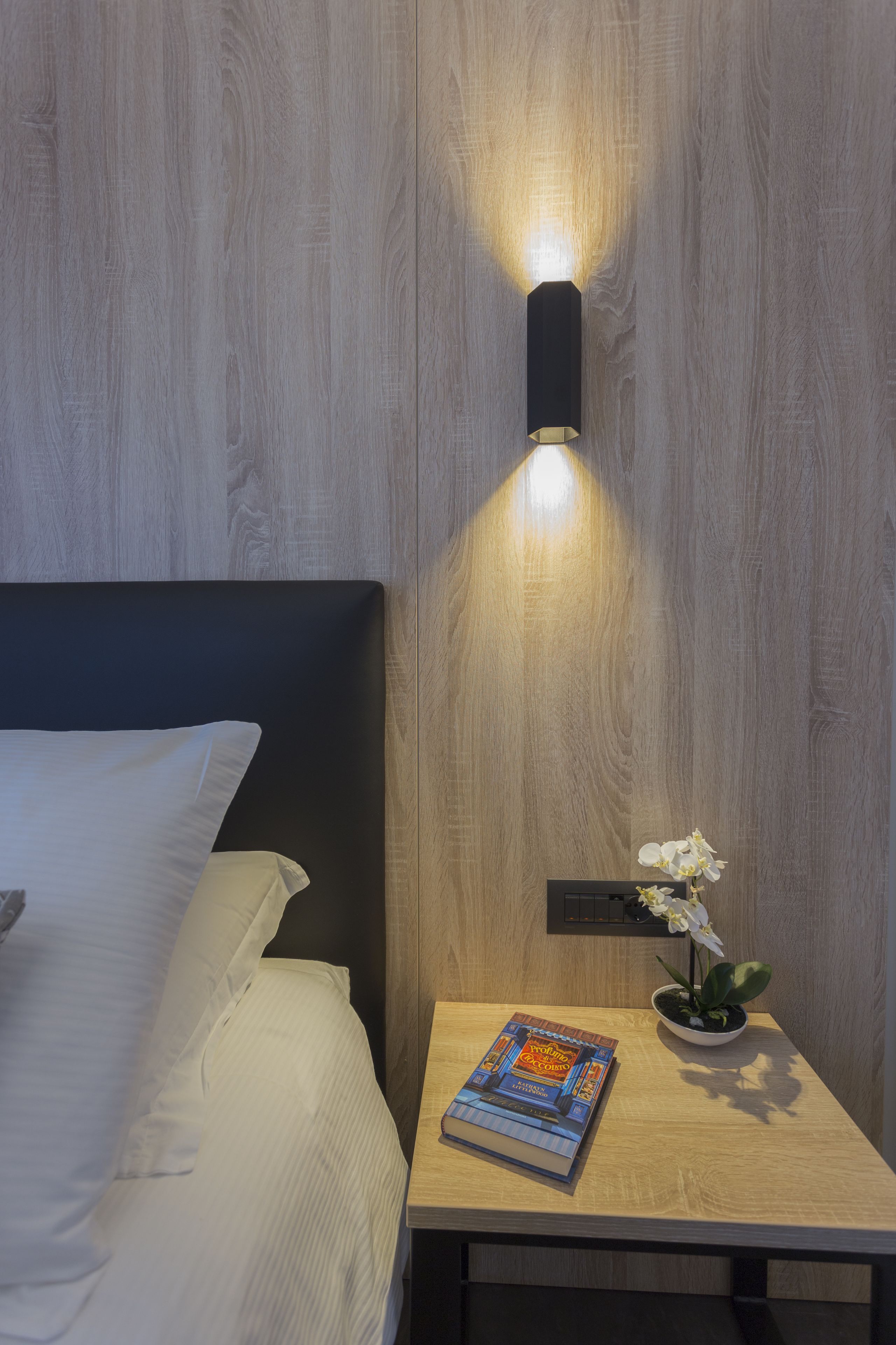 A neatly made bed with white linens, a bedside table with a book and a small potted plant, and a wall-mounted light fixture.