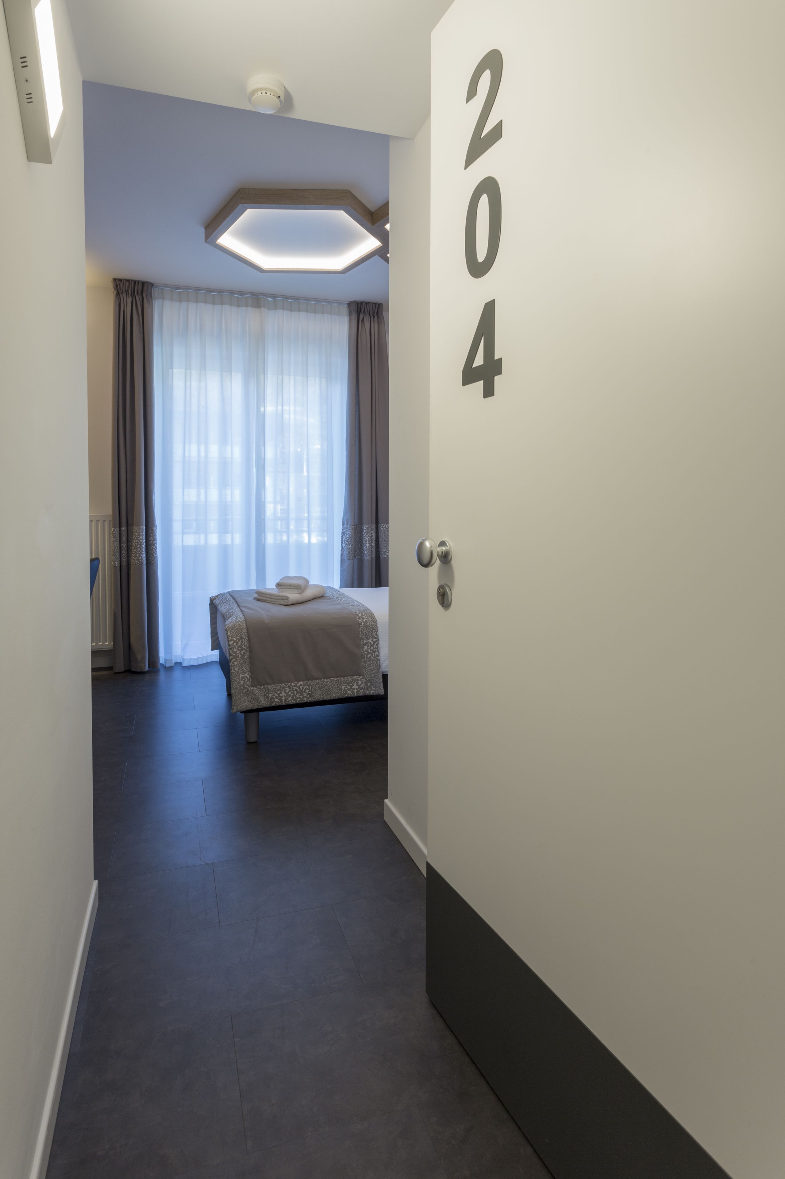 Una habitación de hotel con una cama, el número 204 en la pared, una lámpara en el techo y vistas a través de una ventana con cortinas.
