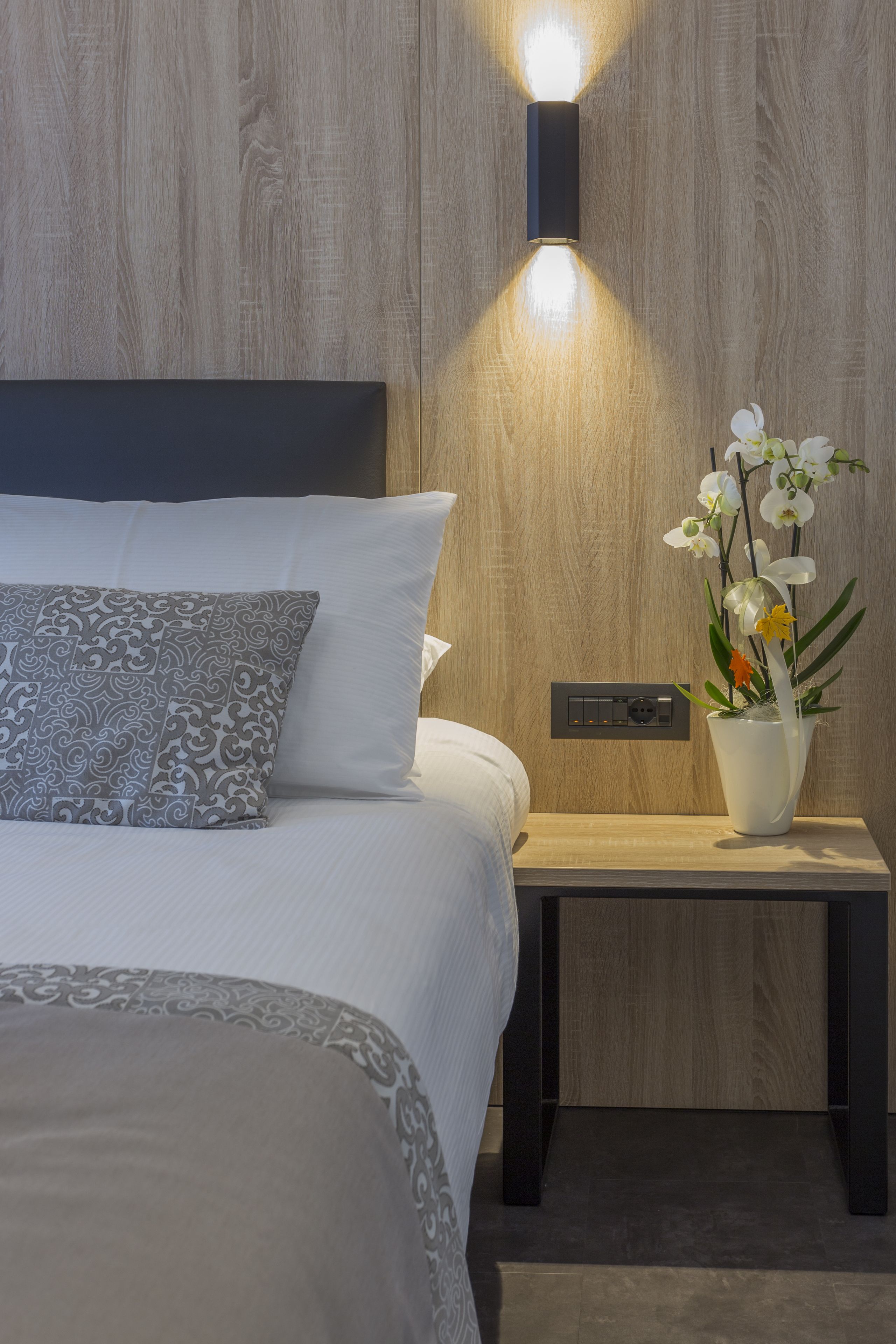 Un lit bien fait, agrémenté d’oreillers à motifs, une table de chevet avec un vase de fleurs et un luminaire fixé au mur.