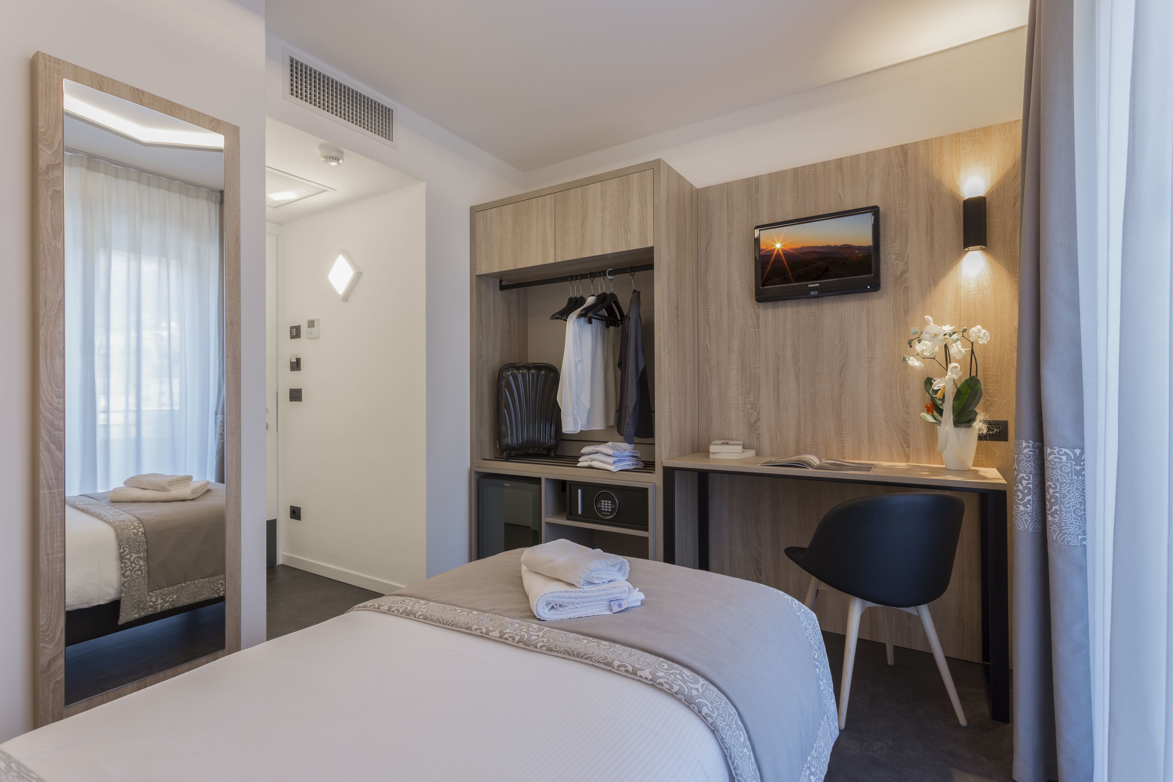 A modern hotel room with a bed, a desk with a chair, a wardrobe, and a TV mounted on the wall.