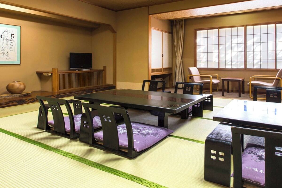 Una habitación japonesa tradicional con suelo de tatami, una mesa baja, sillas, un televisor y una ventana con cortinas.