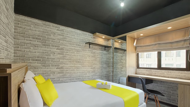 A hotel room with a brick wall, a bed with white and yellow bedding, a desk with a chair, and a window with blinds. - Literooms At Kebagusan City (Jakarta)