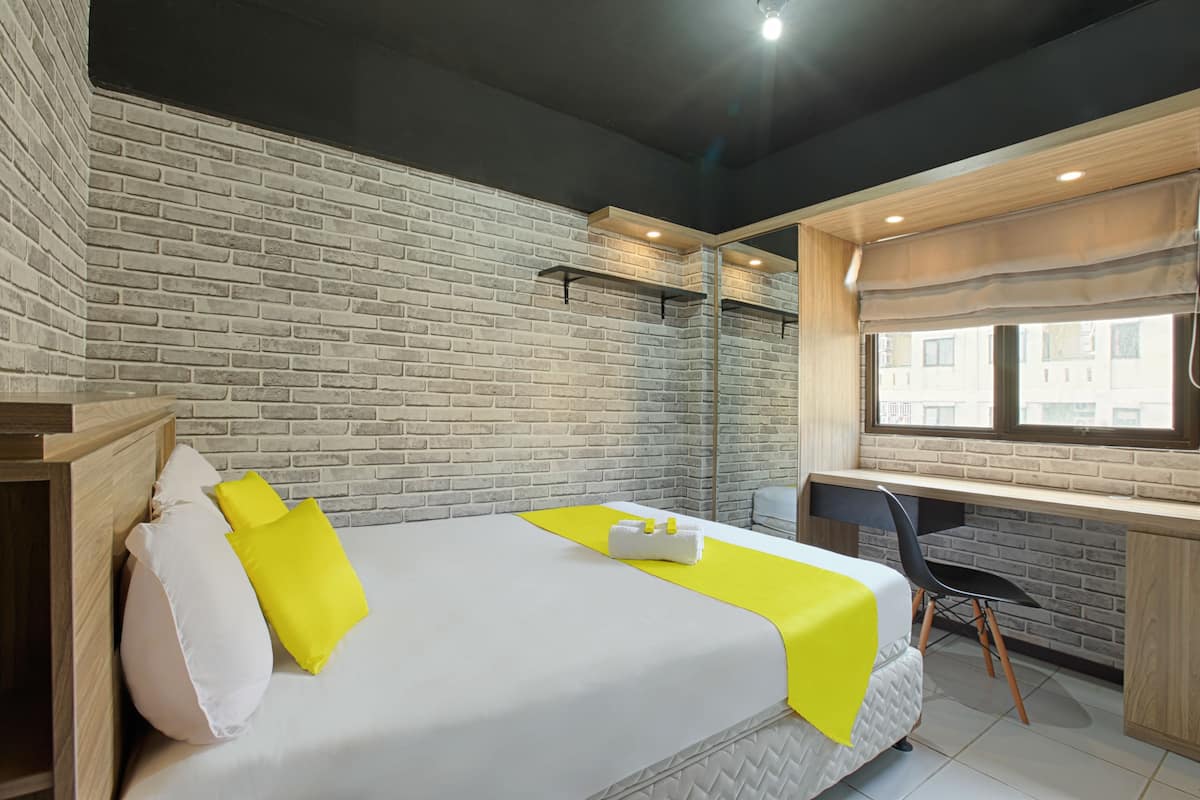 Une chambre d’hôtel avec un mur en briques, un lit avec une literie blanche et jaune, un bureau avec une chaise et une fenêtre avec des stores.