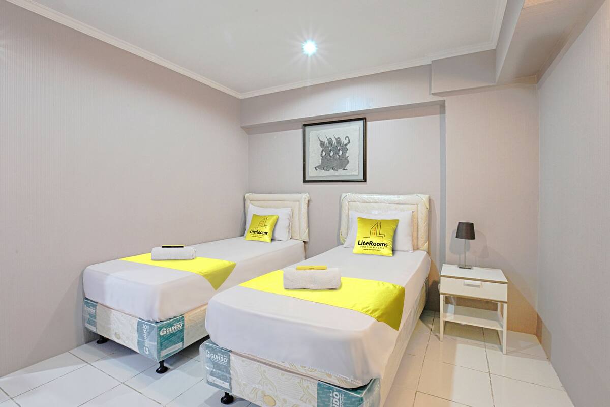 Une chambre d’hôtel avec deux lits, chacun doté d’un matelas blanc et d’une couverture jaune. Un tableau encadré est accroché au mur au-dessus des lits, ainsi qu’une petite table de chevet avec une lampe et une commode blanche.