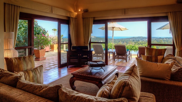 Un salón amplio con un sofá grande, una mesa de centro y un televisor. Hay dos sillones y una mesa con sombrilla. La habitación tiene ventanas grandes con vistas al paisaje.
