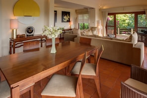 A dining area with a wooden table, chairs, and a vase of flowers. There is a sofa and a window with a view of greenery. - Alpha Villa (Koh Samui)