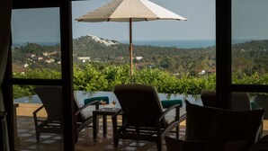 Habitación con vistas a una colina y al mar. Hay dos sillas y una mesa con una bebida.