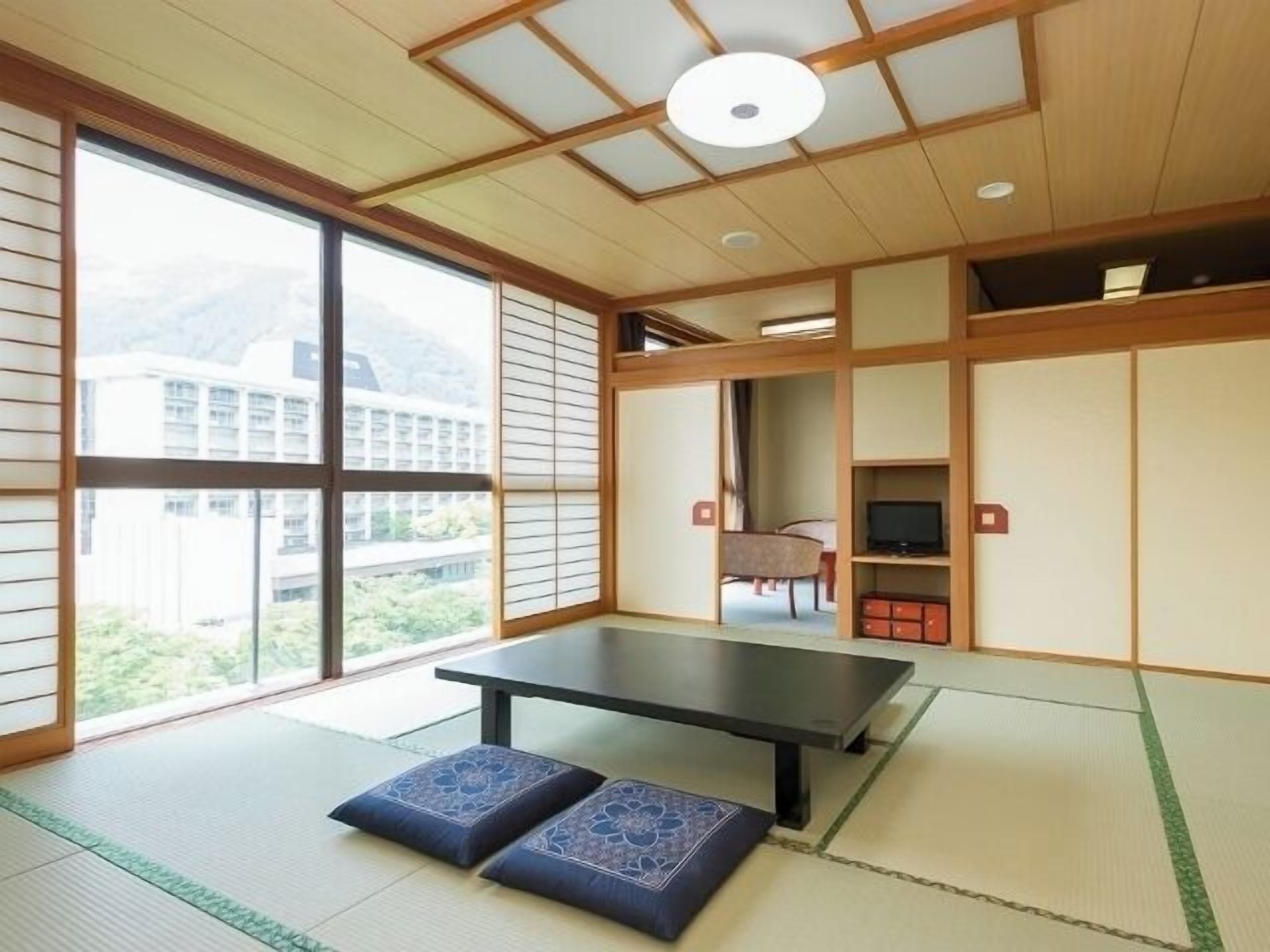 A tatami mat room with a tatami mat table, two cushions, a sliding door, and a view of a building outside.