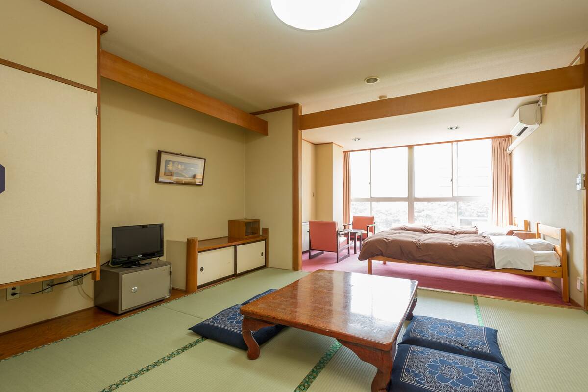 Ett traditionellt japanskt rum med tatamimattor på golvet, en låg säng av trä, en tv-apparat placerad på ett skåp och en skjutdörr som leder ut till en balkong.