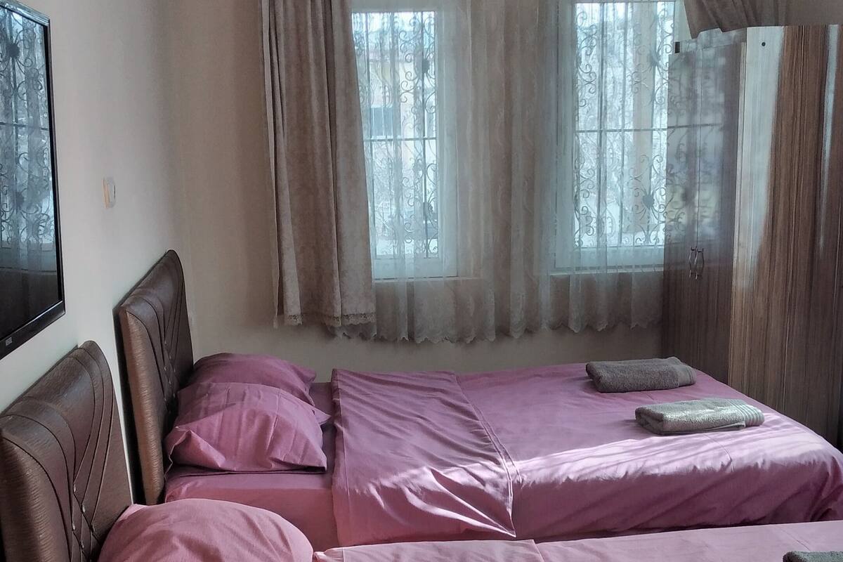A bedroom with a large bed, two windows with curtains, a wooden headboard, and a ceiling light.