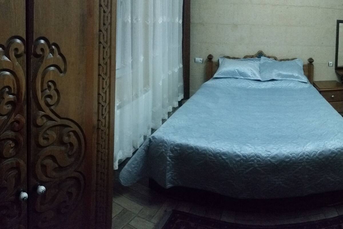 A bedroom with a bed, a wooden wardrobe, a small bedside table, a chair, a television, and a mirror on the wall.