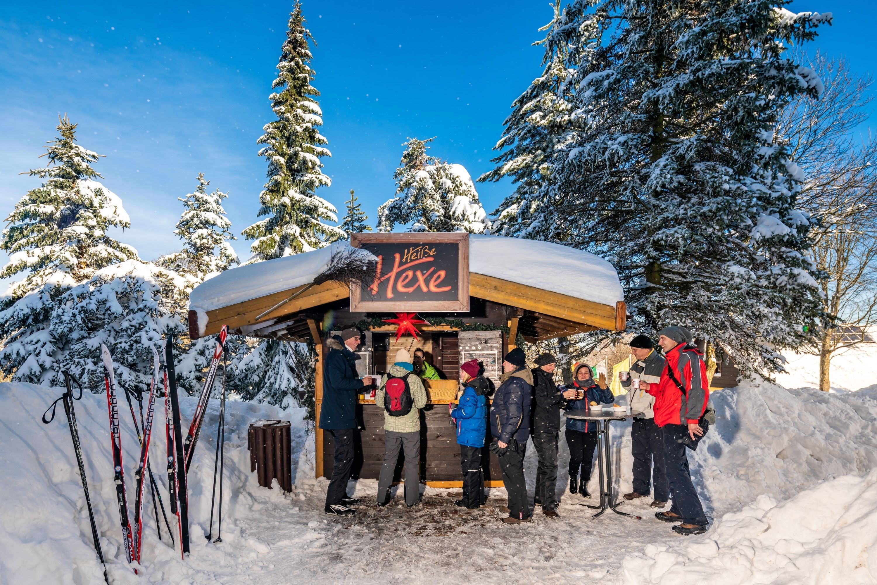 Een besneeuwde buitenruimte met een houten kiosk, genaamd 'Hütte Hexe', en verschillende mensen eromheen.