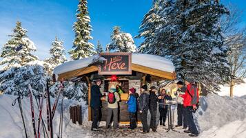 Een besneeuwde buitenruimte met een houten kiosk, genaamd 'Hütte Hexe', en verschillende mensen eromheen.