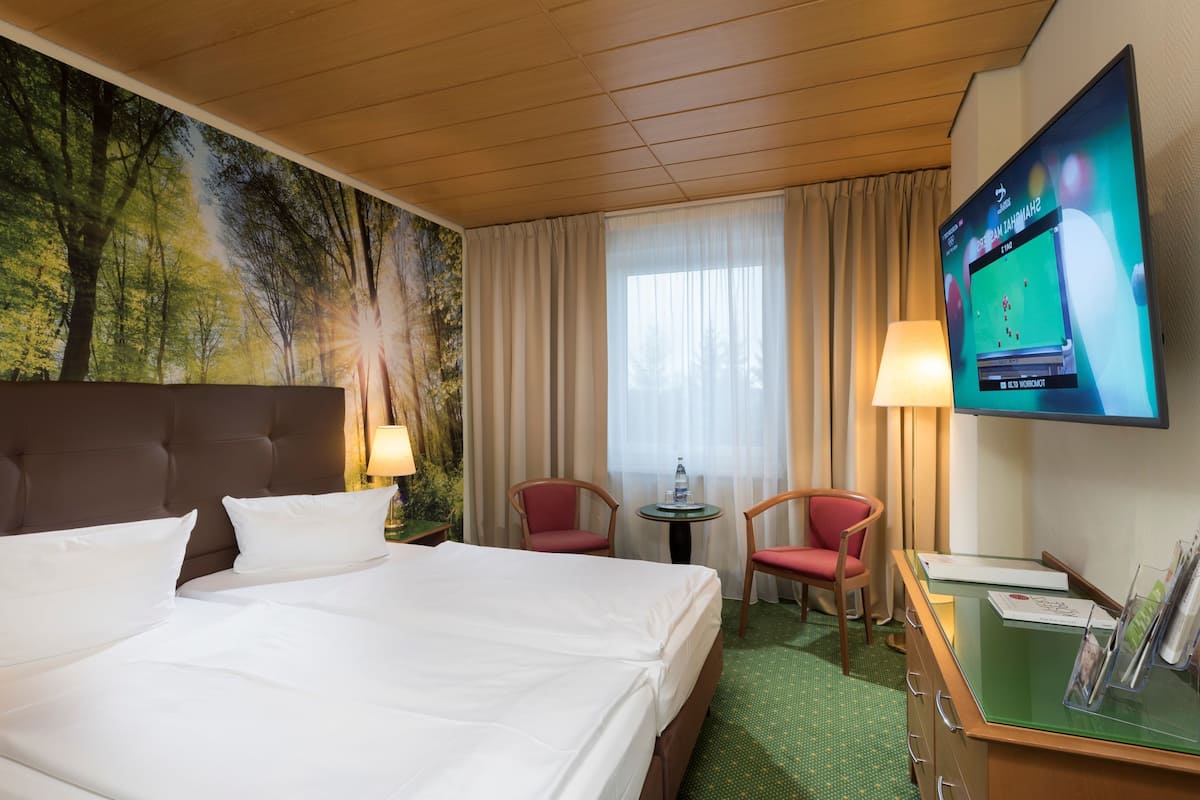 A hotel room with a bed, two chairs, a desk, a television, and a mural of trees.