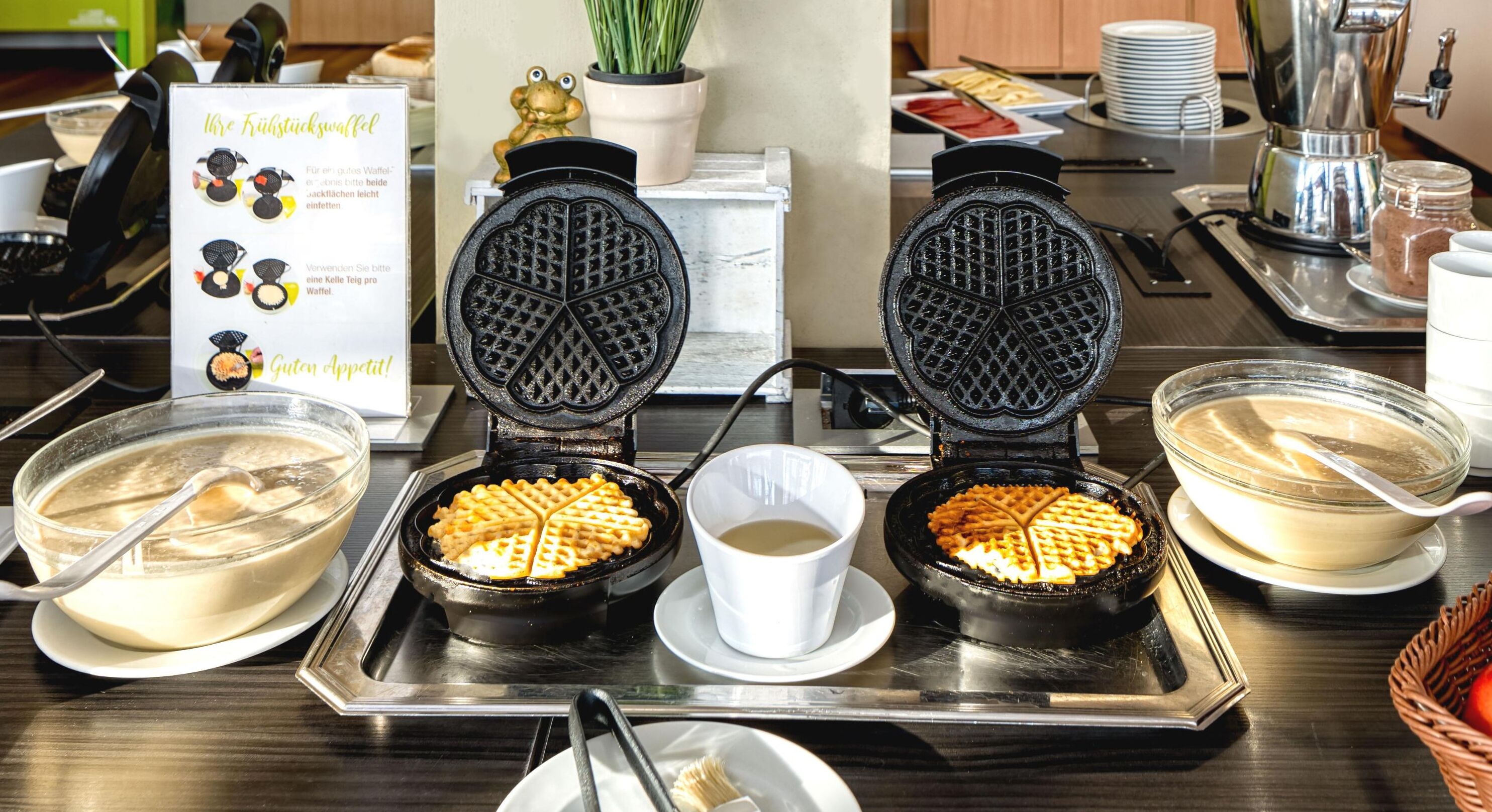 A waffle station with two waffle irons, a bowl of batter, a cup of syrup, and a sign with waffle recipes.