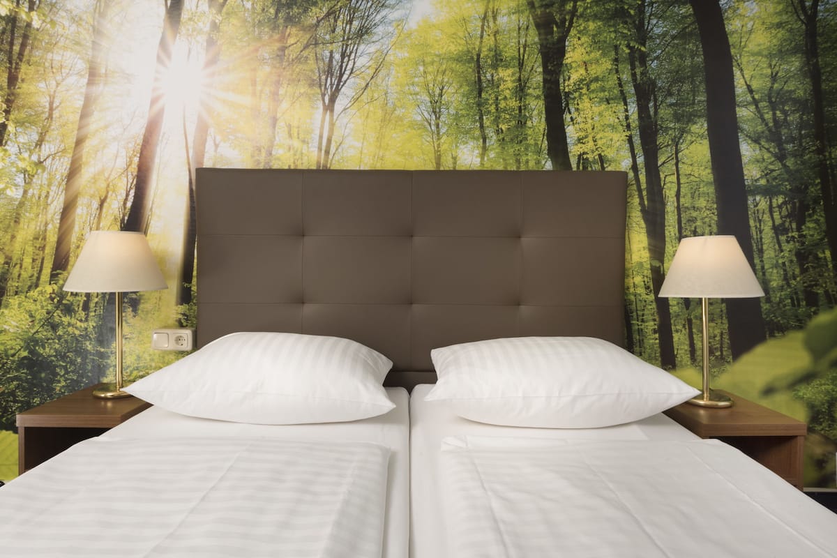 A double bed with a tufted headboard, two bedside lamps, and a forest-themed wall mural.