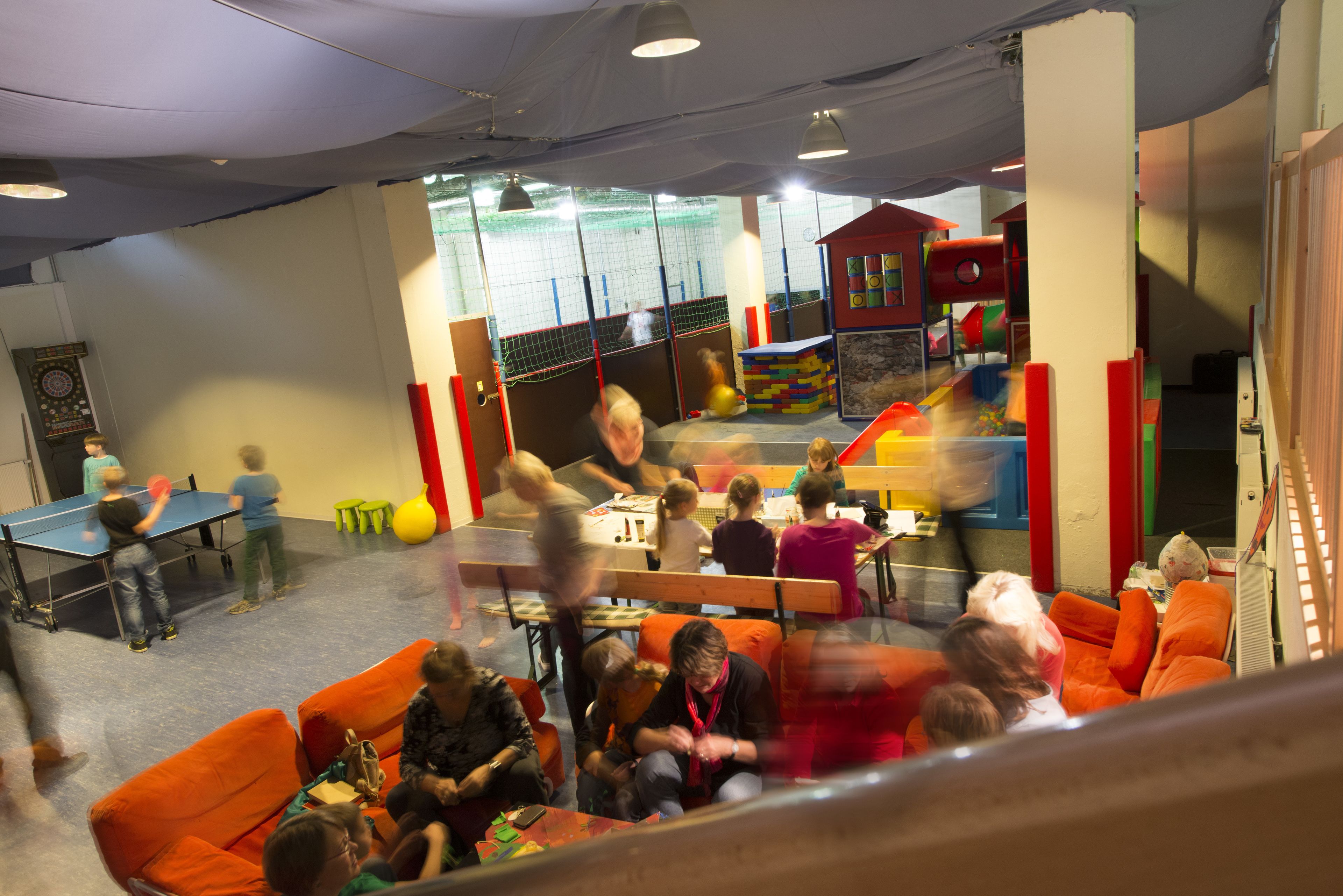 Une pièce équipée d’une table de ping-pong, de sièges orange et d’un espace de jeux avec des structures colorées.