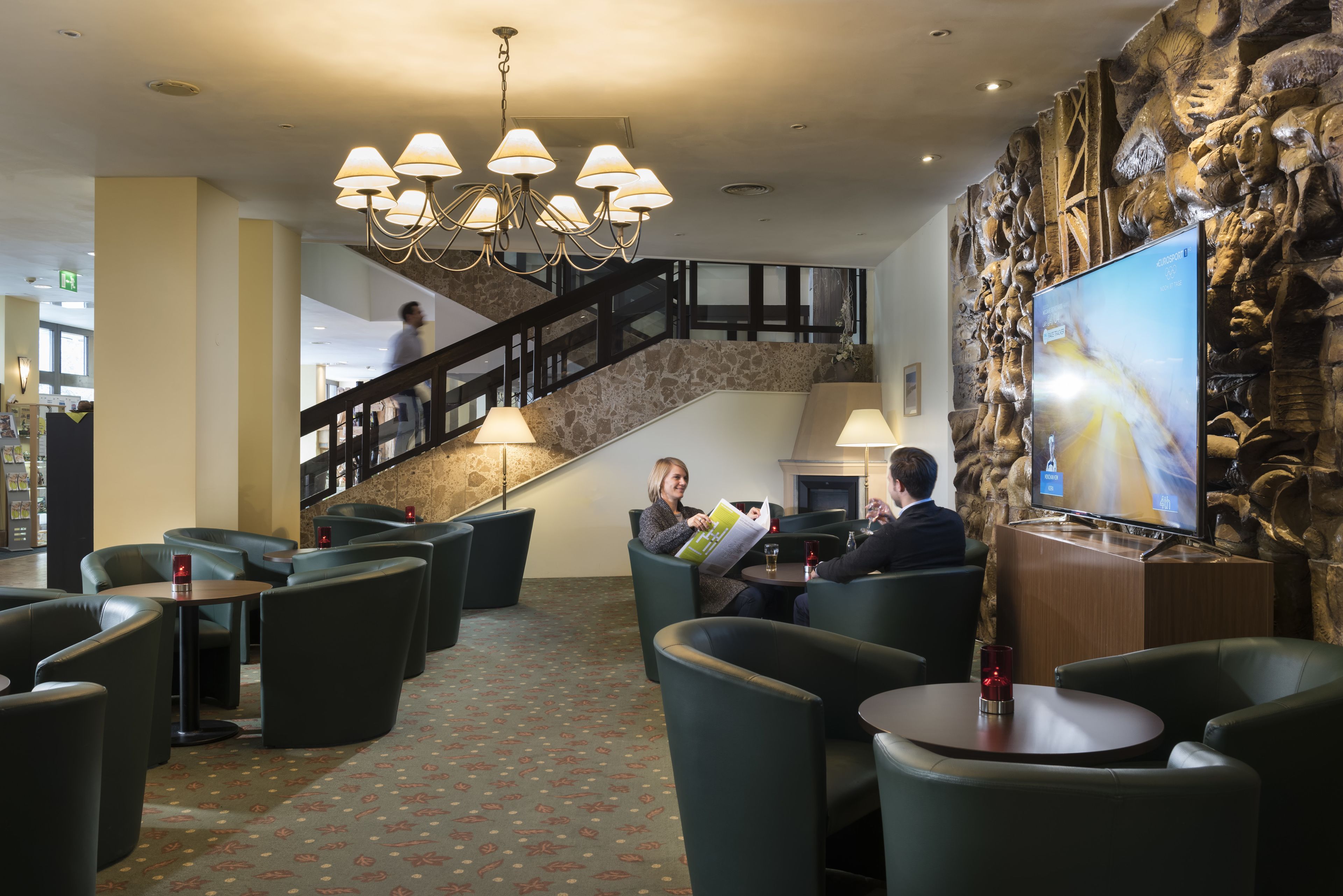 A lobby with a staircase, seating areas, a TV screen, and a chandelier.