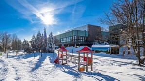A snow-covered playground with red structures, surrounded by trees and a building with large windows. - AHORN Waldhotel Altenberg (Altenberg)