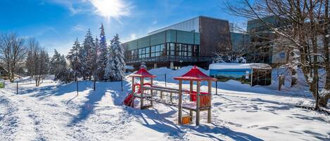 A snow-covered playground with red structures, surrounded by trees and a building with large windows.