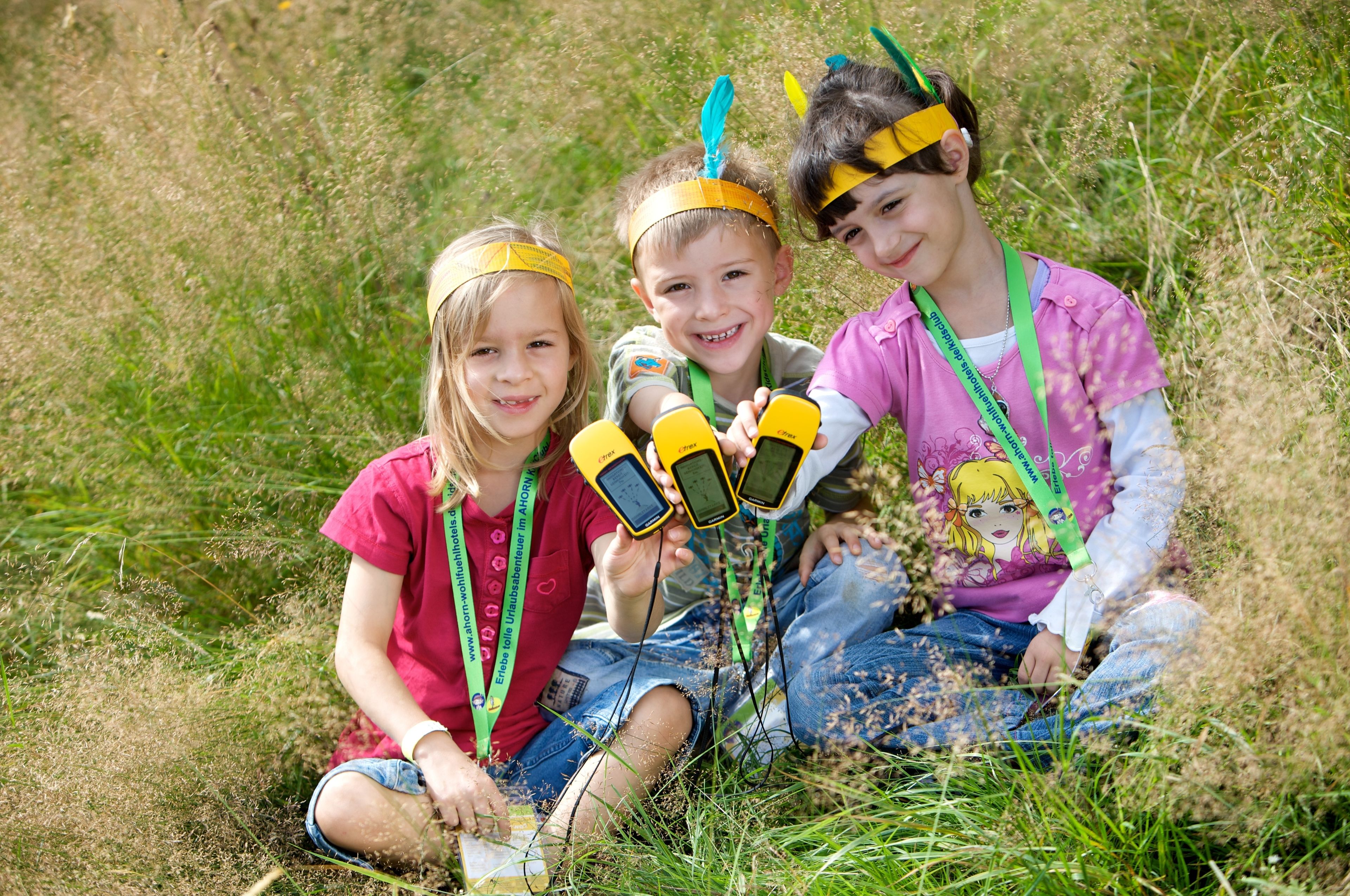 Three children sitting on grass, wearing headbands and holding yellow devices.