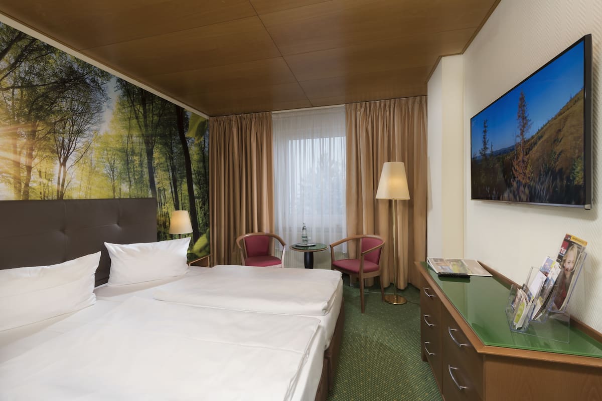 A hotel room with a large bed, two chairs, a small table, a lamp, a desk with a magazine rack, and a painting on the wall.