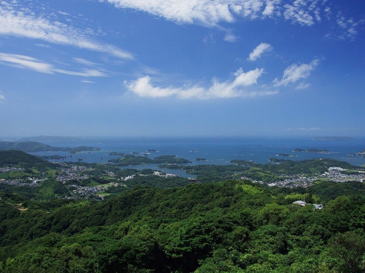 A coastal landscape with dense greenery, a clear sky, and a view of the sea and distant islands.