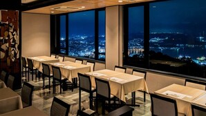 A dining area with tables and chairs, large windows with a city view at night.