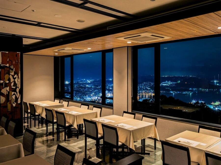A dining area with tables and chairs, large windows with a city view at night.