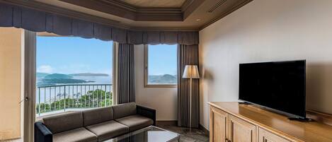A hotel room with a sofa, a TV, a glass coffee table, and a view of the sea.