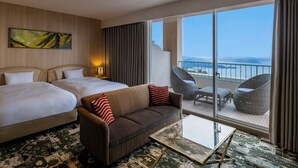 A hotel room with two beds, a sofa, a balcony with wicker chairs, and a large window with a view of the sea.