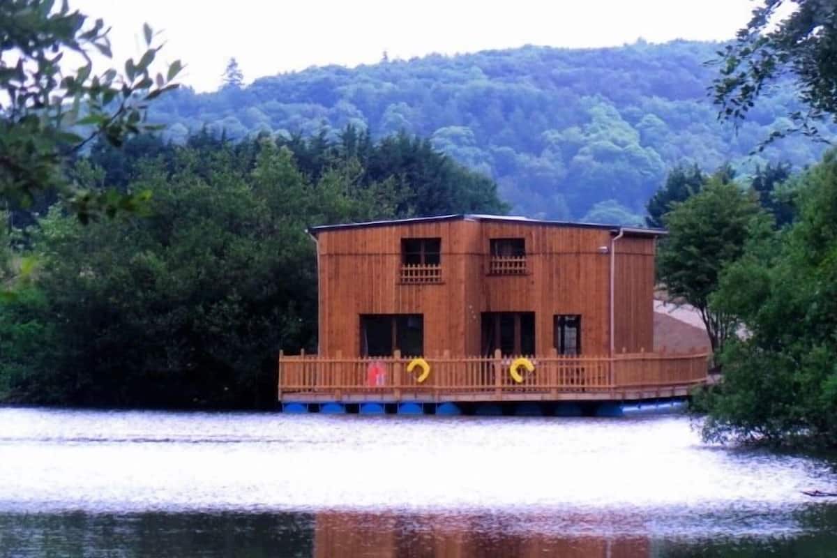 A wooden cabin with a balcony, situated on a lake surrounded by trees.