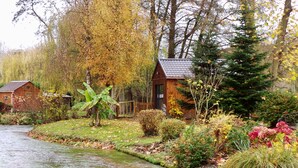 A riverside property with a wooden cabin, a small shed, and a well-maintained garden with various plants and a banana tree. - Au Fil de L'eau (Colleville)